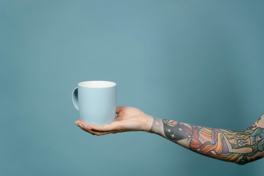 A tattooed arm holds a light blue mug against a solid blue background in a studio setting.